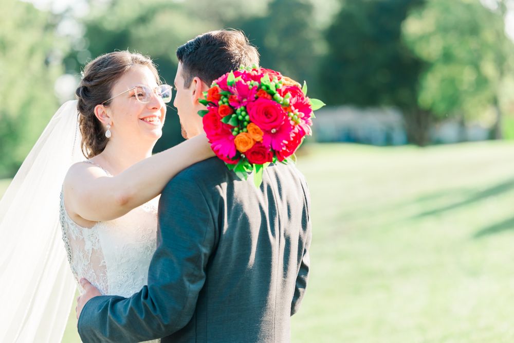 Wedding couple embraces while holding vibrant red bouquet against outdoor garden setting.