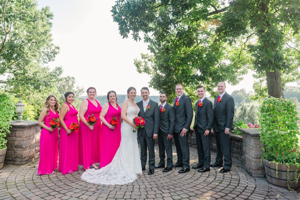 Wedding party in black suits and bright pink dresses poses on stone patio with landscaped background.