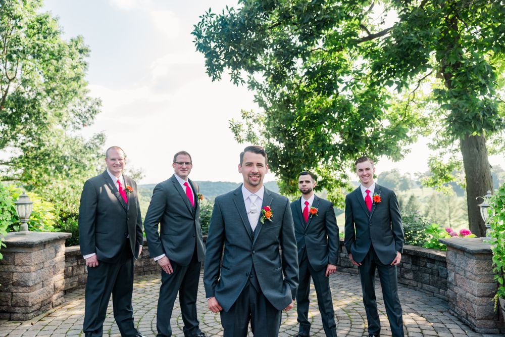 Wedding party of groomsmen in dark suits pose on stone terrace with trees in background.