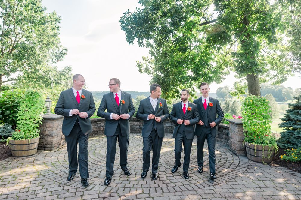 Groomsmen in black suits with red ties stand together on a stone patio under trees.