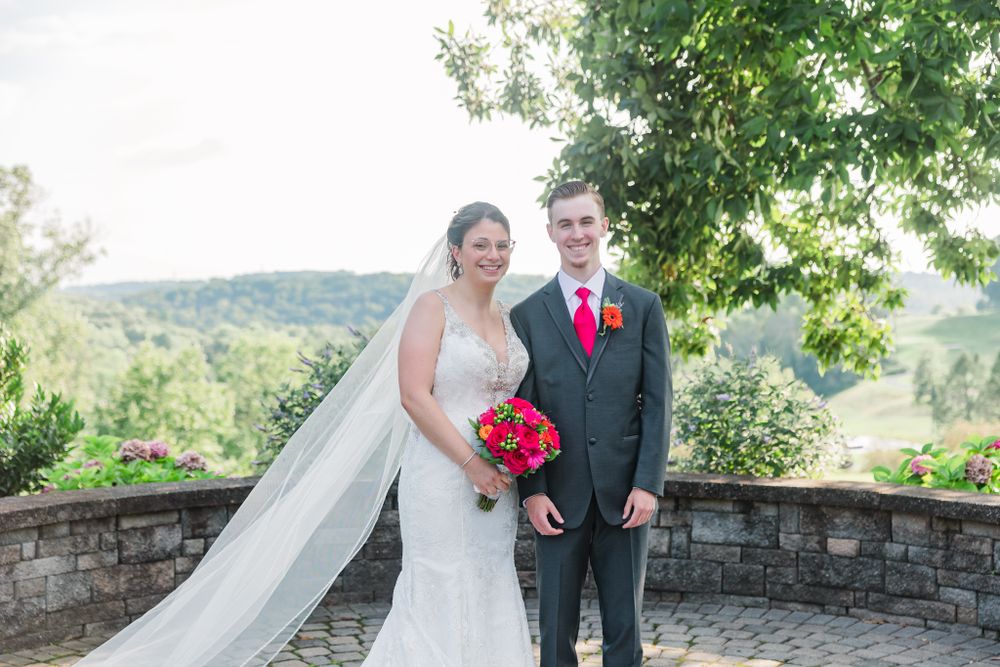 A newlywed couple poses on a stone terrace overlooking scenic hills with the bride holding a vibrant red bouquet.