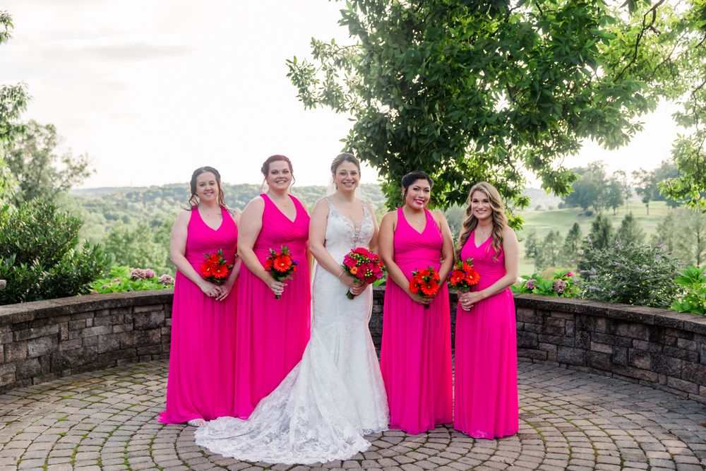 Wedding party posing on stone patio with bridesmaids in bright pink dresses holding red bouquets.