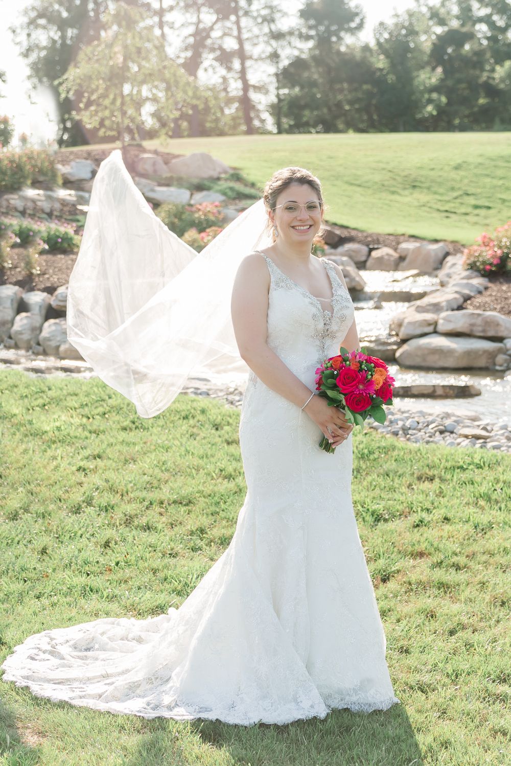 A bride in a flowing white lace gown with a long veil holds a vibrant red bouquet in an outdoor garden setting.