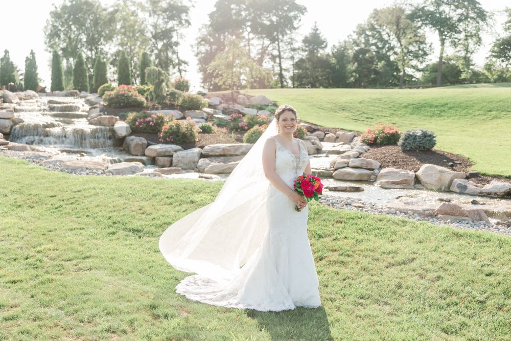 Bride in flowing veil holds red bouquet while twirling on grassy hill with stone waterfall behind.