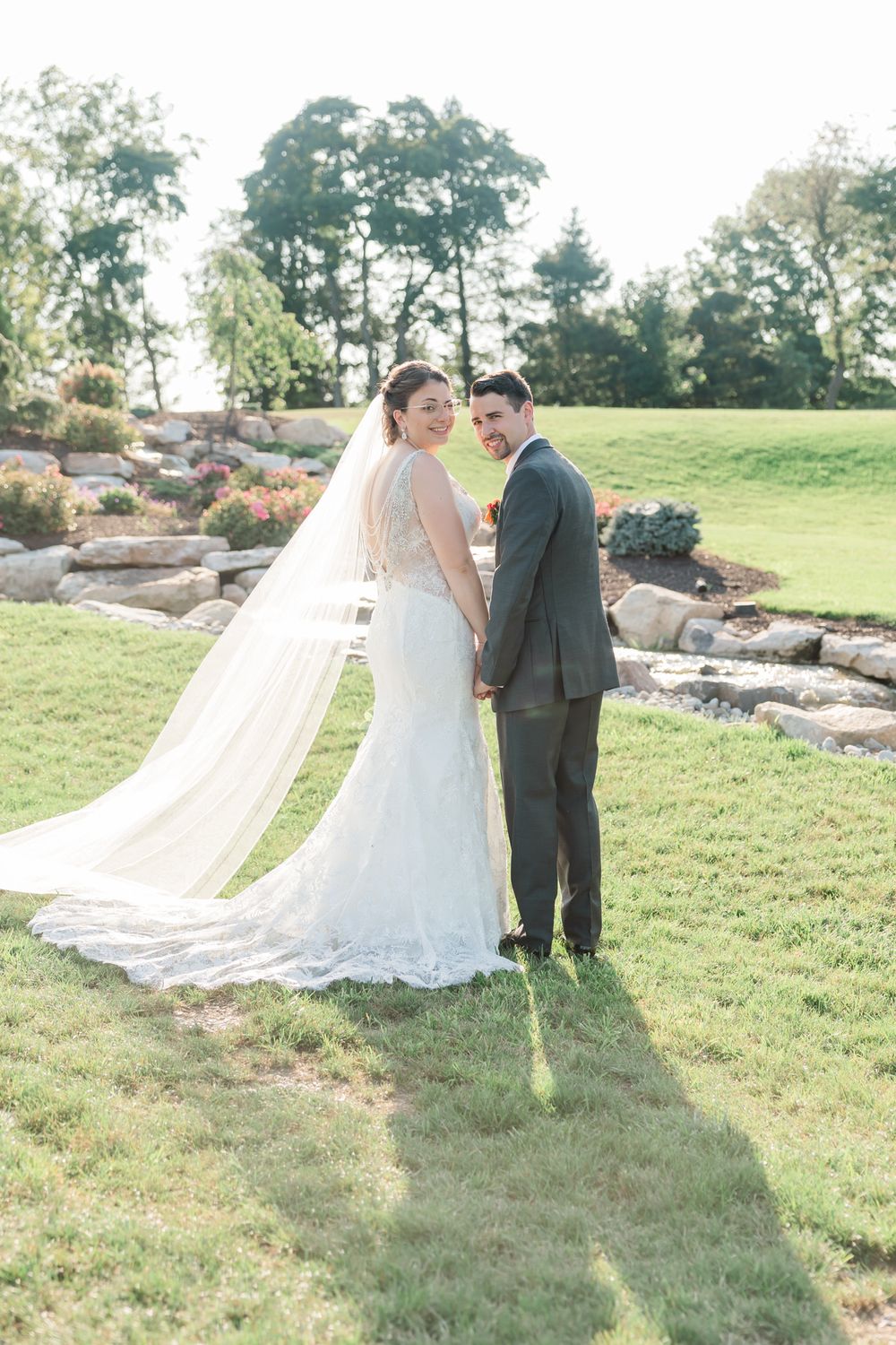 Wedding couple stands together on grassy hill with flowing veil and rock garden in background.