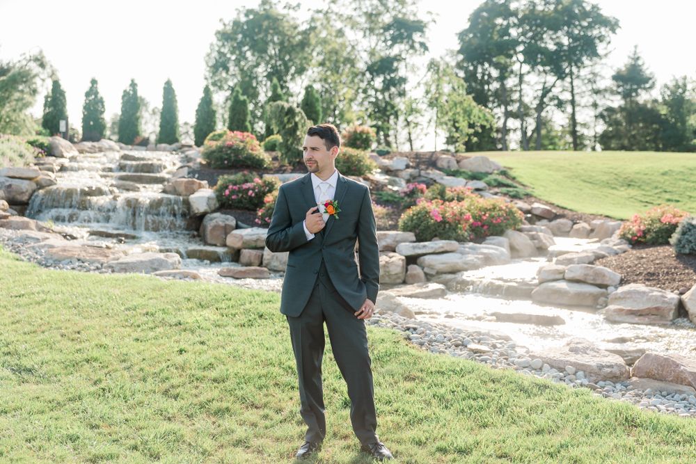 Groom in dark suit stands near outdoor waterfall feature on manicured lawn.