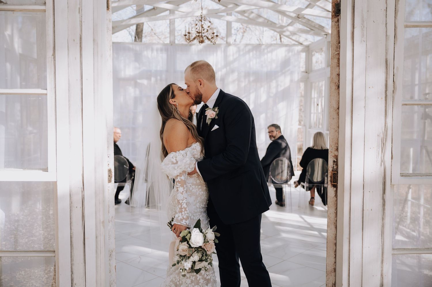 Romantic wedding moment captured in a sun-filled glass chapel with guests in background.
