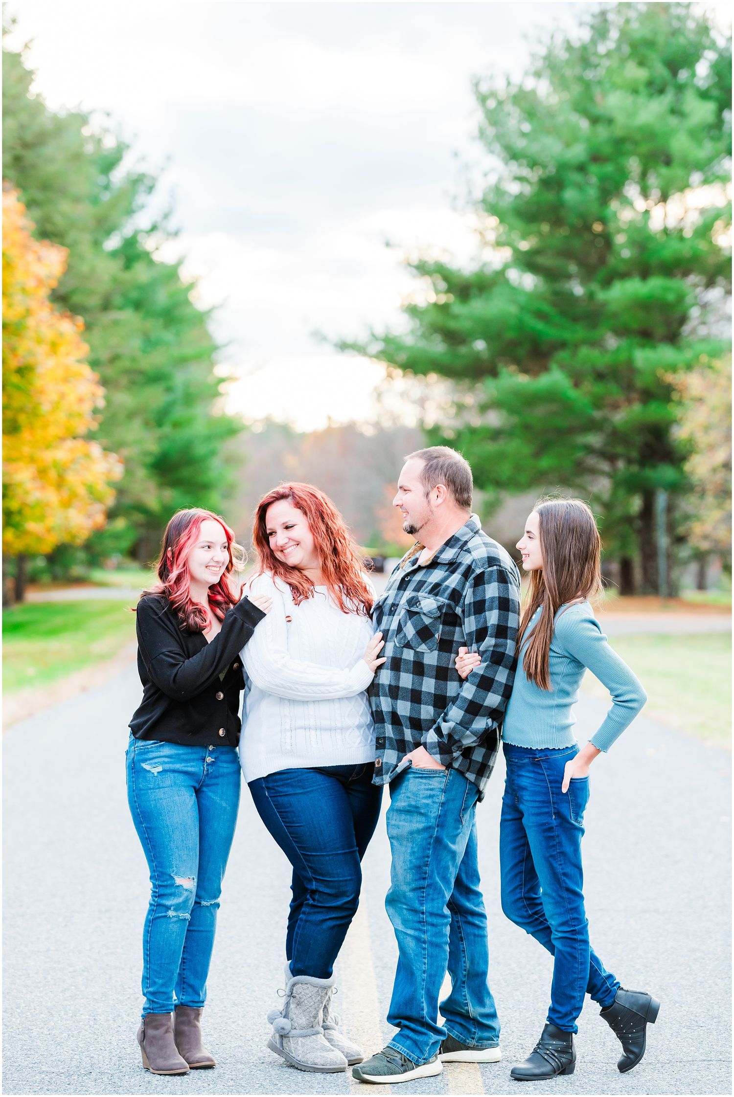 Group of four people in casual attire pose together on a road with fall foliage and pine trees.