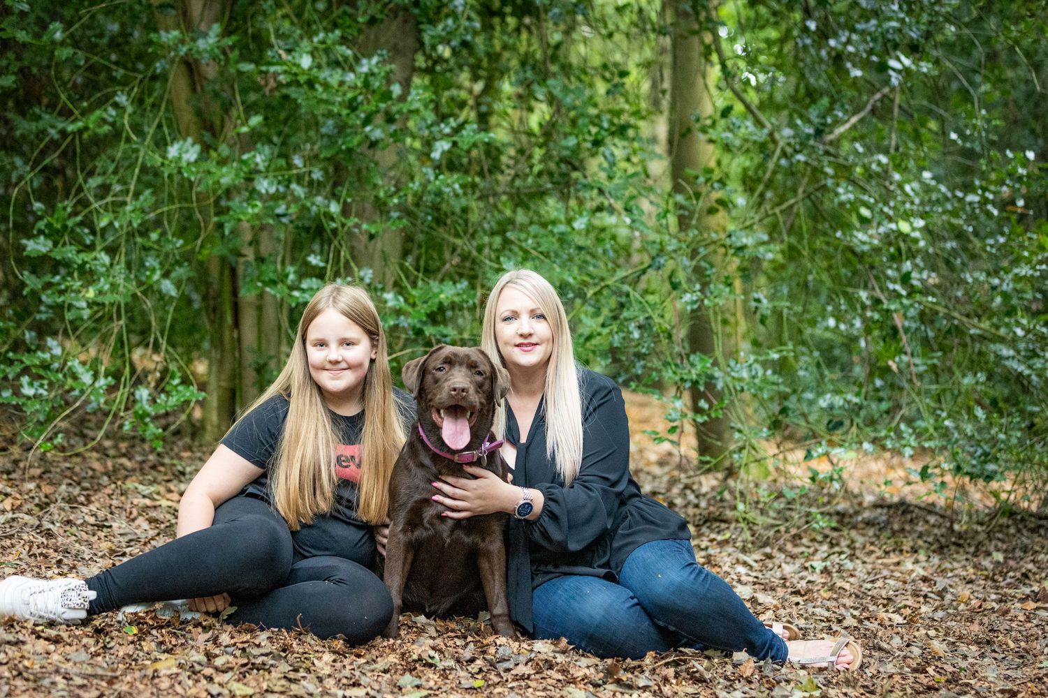 Two girls and their chocolate lab dog pose together in a wooded outdoor setting.