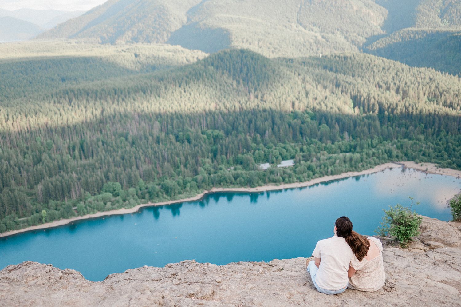 A couple sits on a mountain cliff overlooking a blue lake below