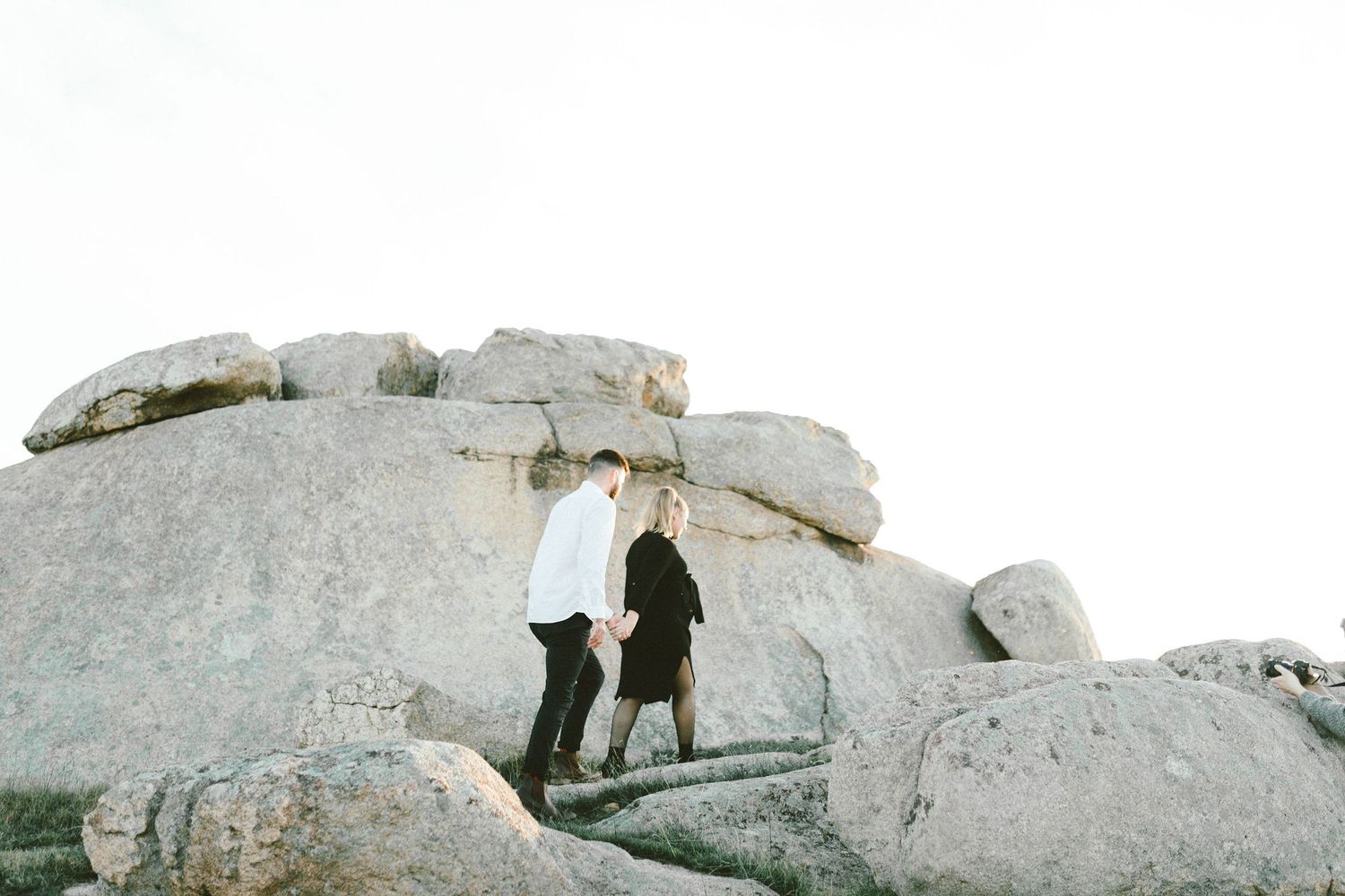 Two silhouettes stand together on a rocky hilltop against a bright sky.