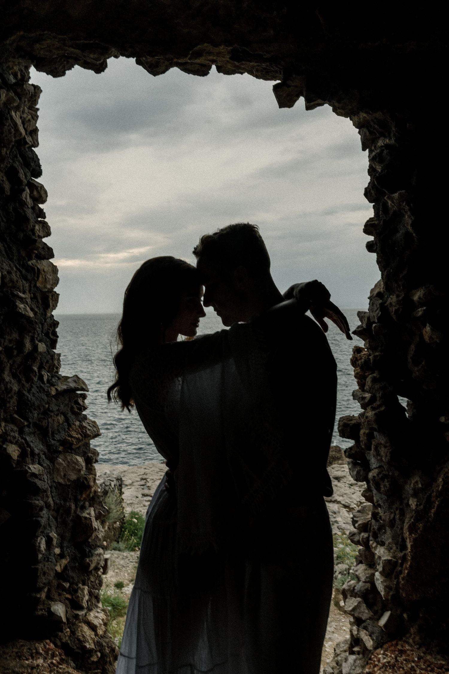A silhouetted couple shares an intimate moment framed by a stone archway overlooking the ocean.