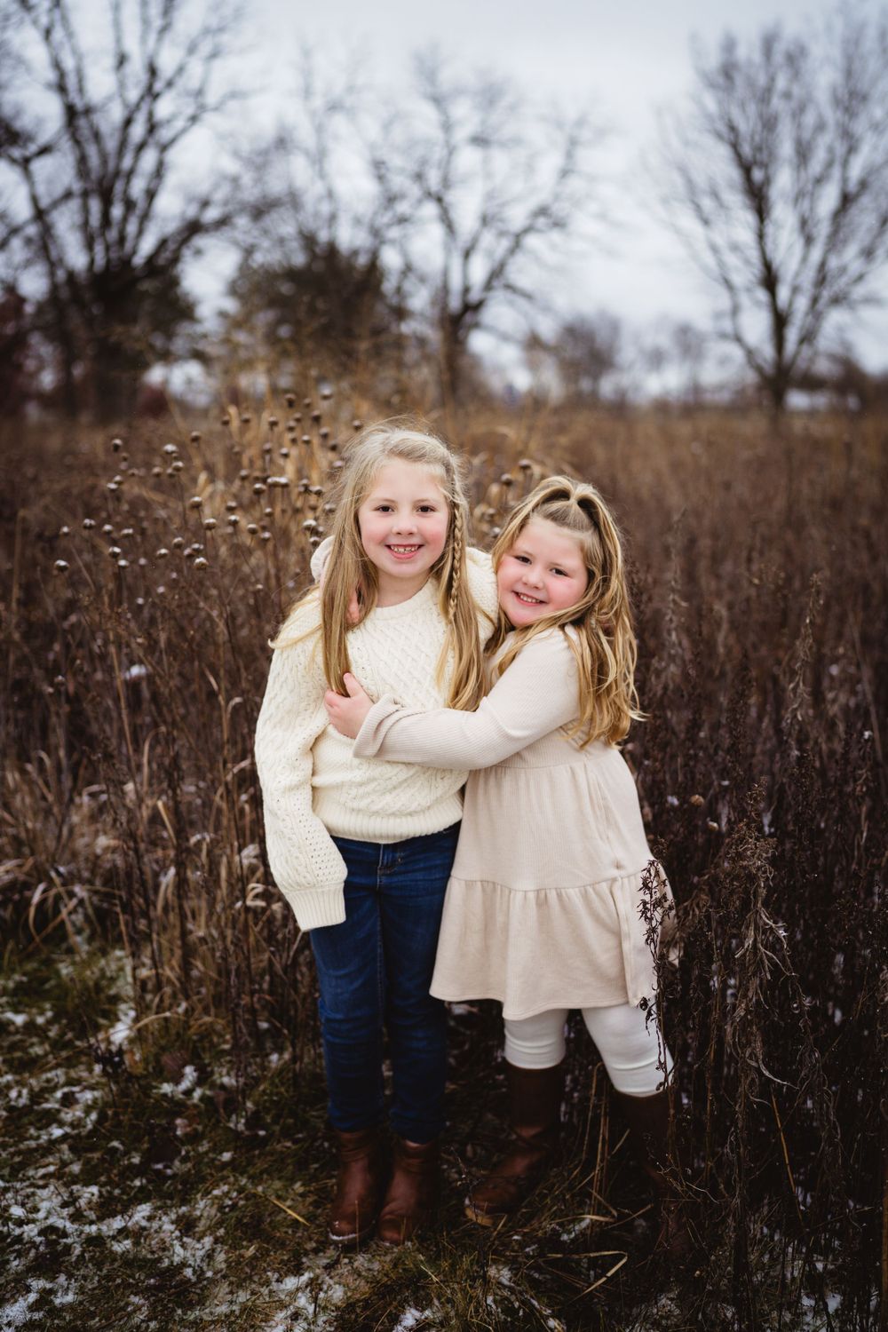 Two young girls in cream-colored outfits hugging and smiling in a winter field.