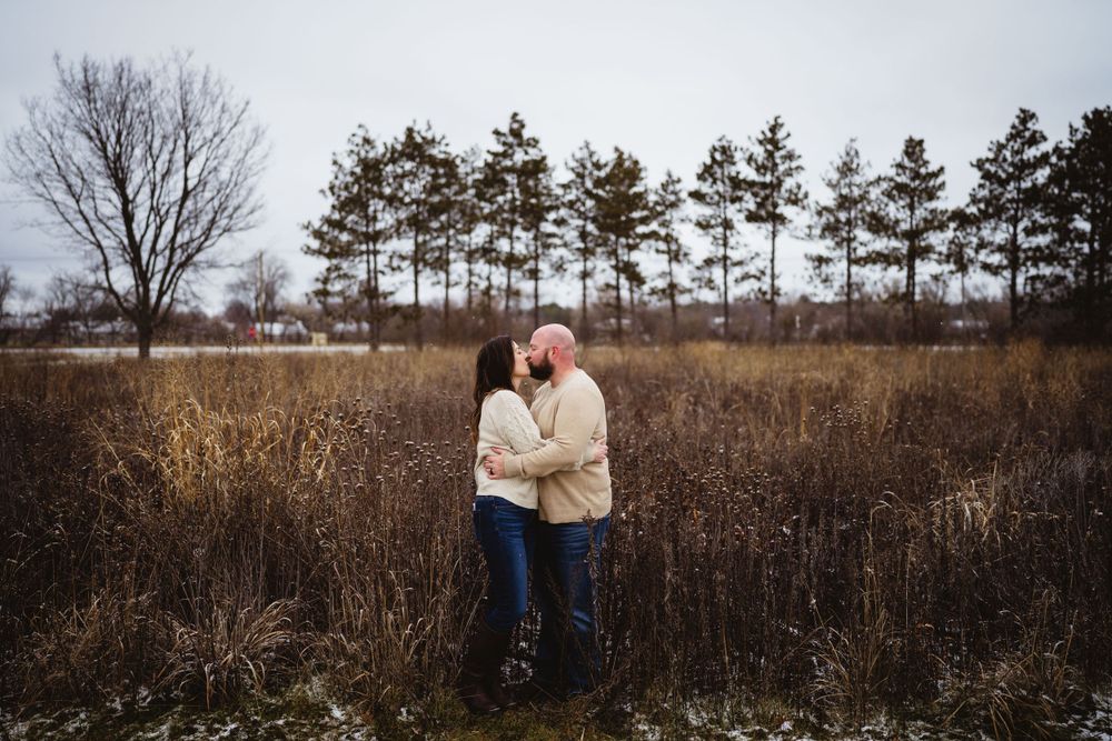 Couple embracing in a winter field with a row of pine trees in the background.