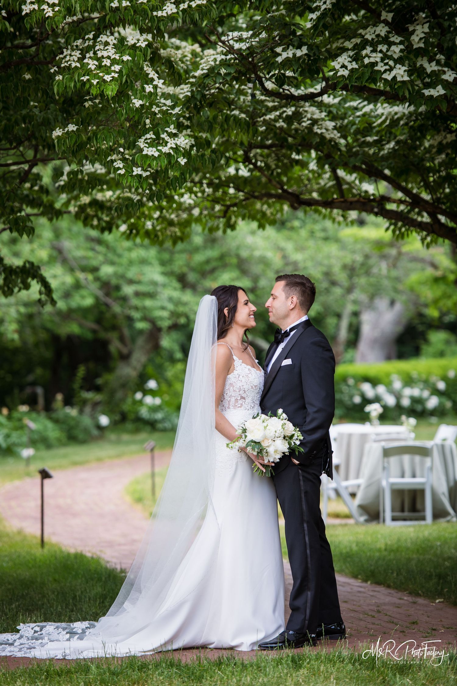Bride and groom gazing at each other on a garden path beneath a flowering tree on their wedding day.