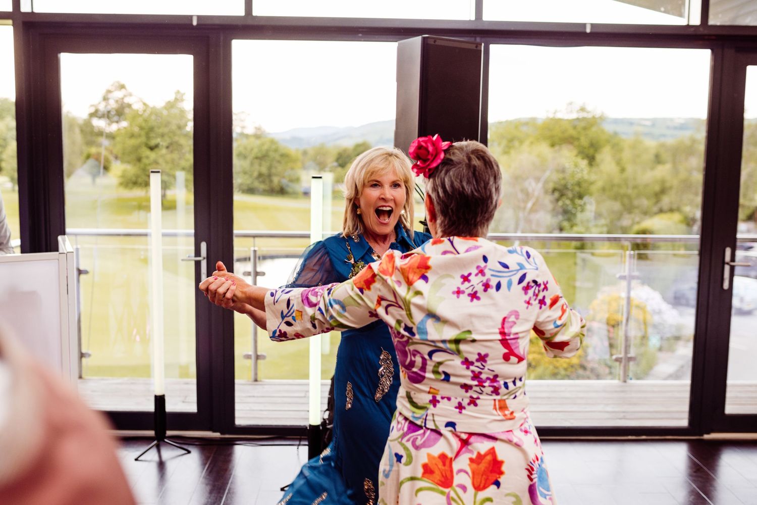 Two wedding guests in colorful attire dancing joyfully by large windows at a reception.