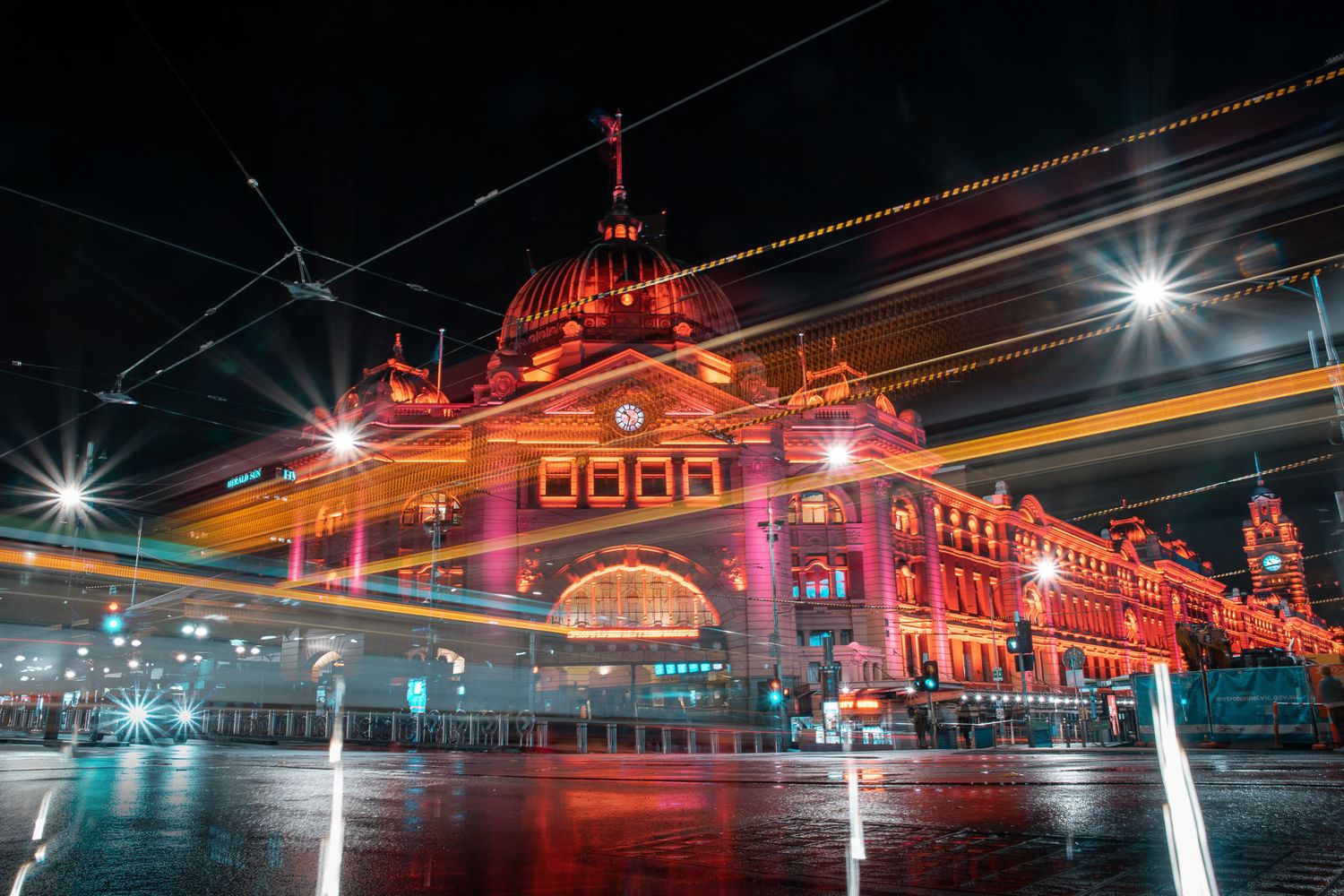 Flinders Street Station in Melbourne lit up at night with light trails and colorful illumination.