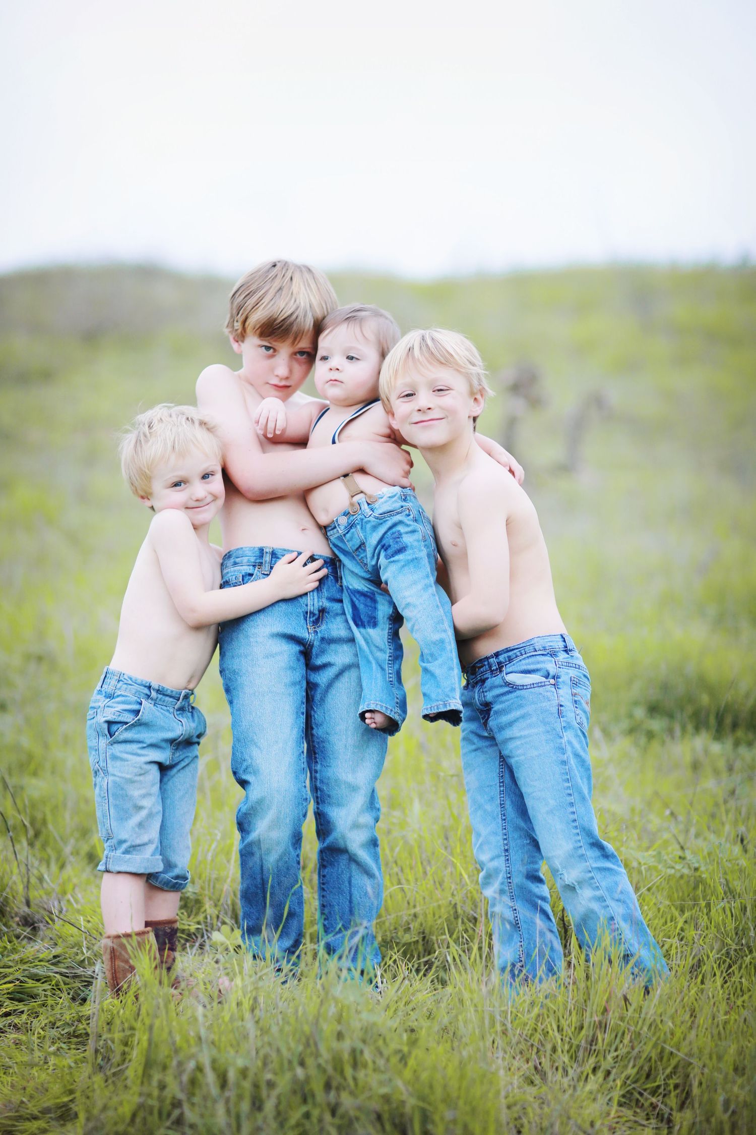Four children in blue jeans embrace together while standing in a grassy field on a sunny day.