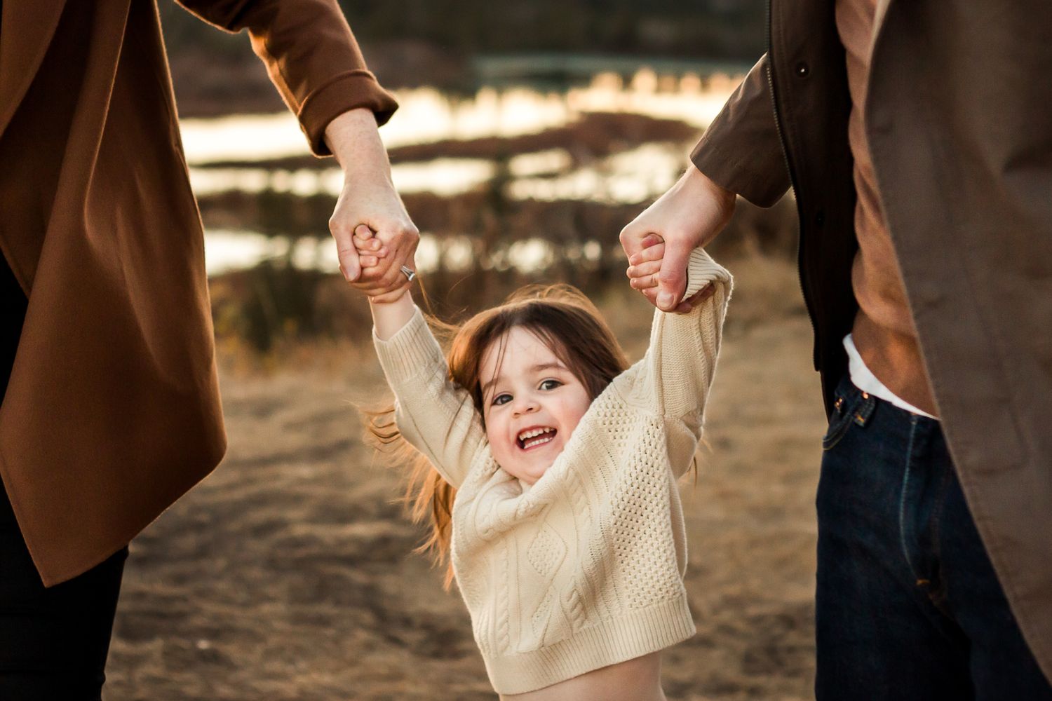 Little girl happily holds parent's hands during fall photos at golden hour at Glenmore Reservoir.