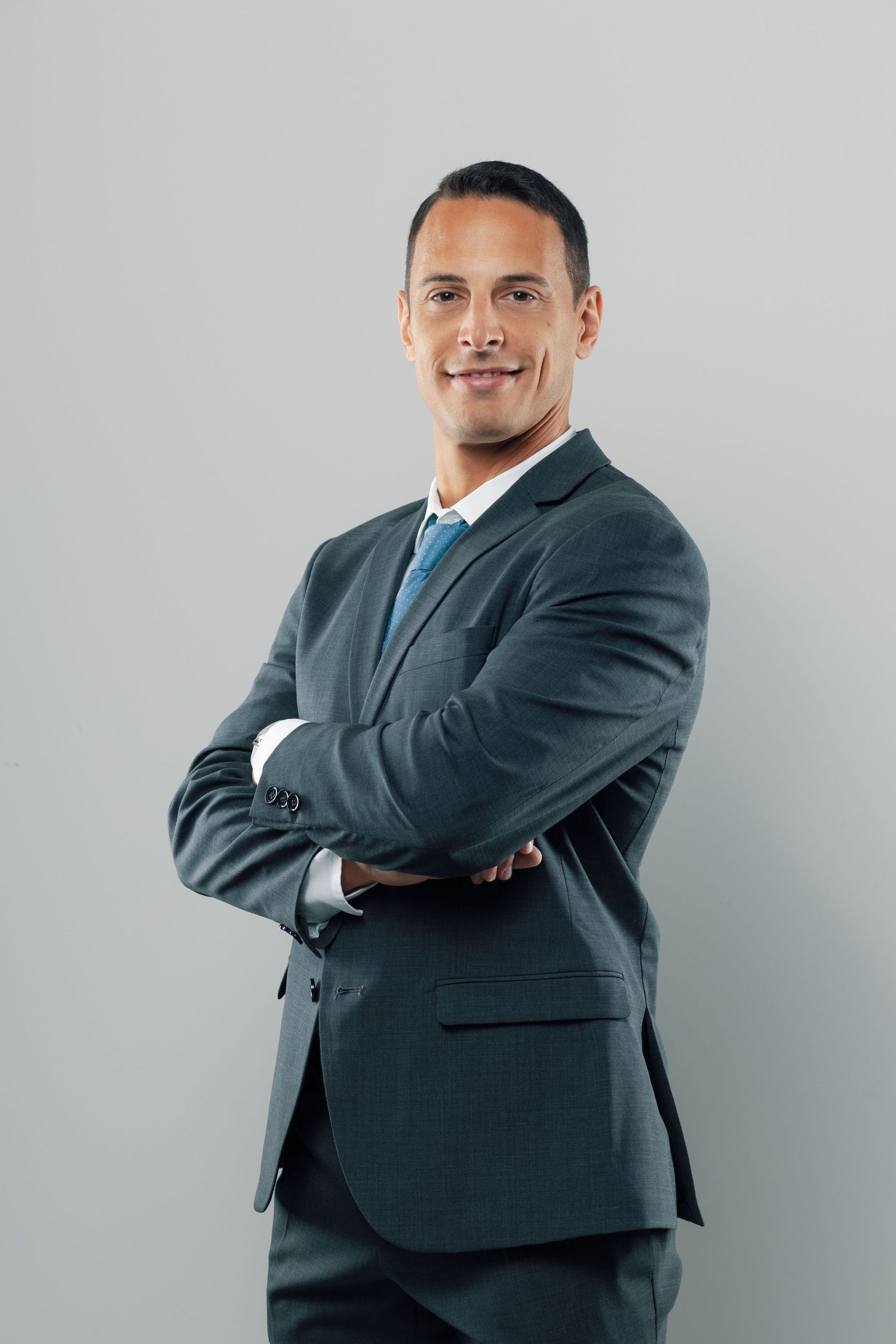 Professional in dark suit and blue tie poses with crossed arms against gray background.