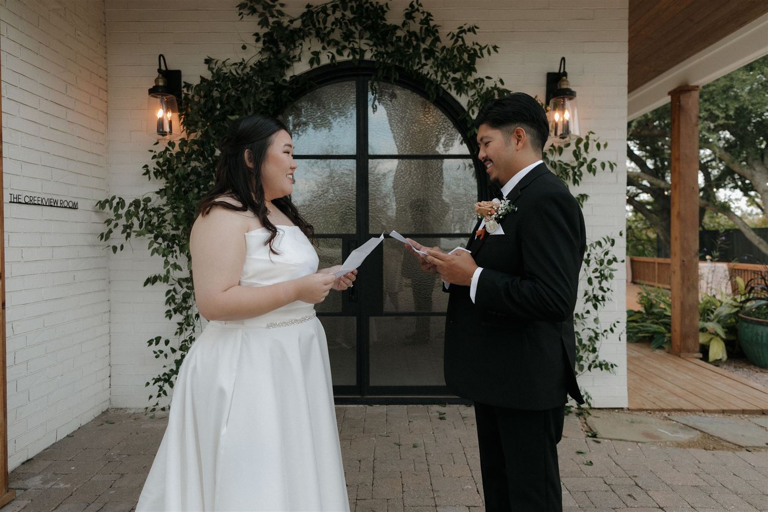 Couple reading their wedding vows under a greenery-decorated archway at white brick venue.
