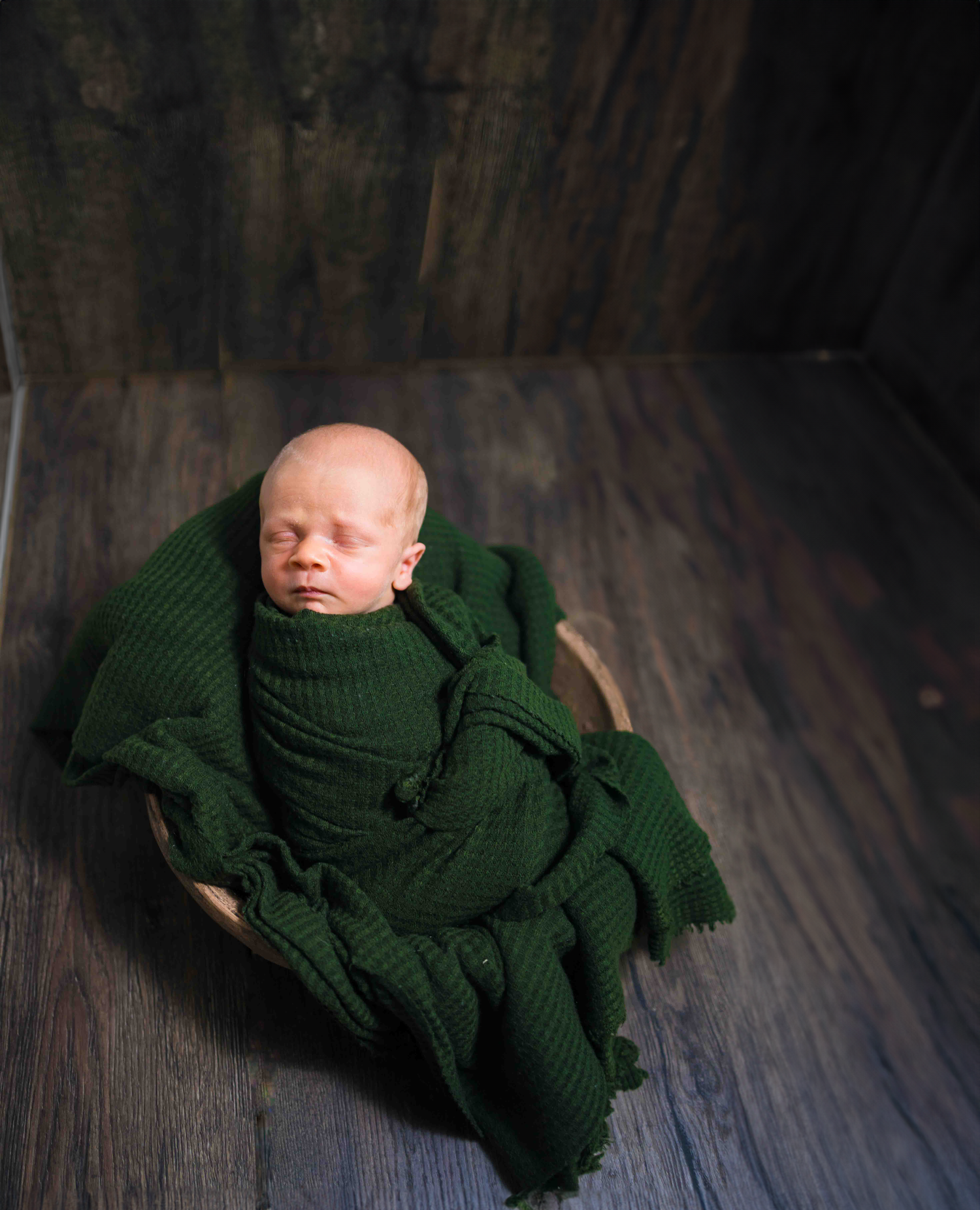 Newborn baby wrapped in green knit blanket on dark wooden floor.