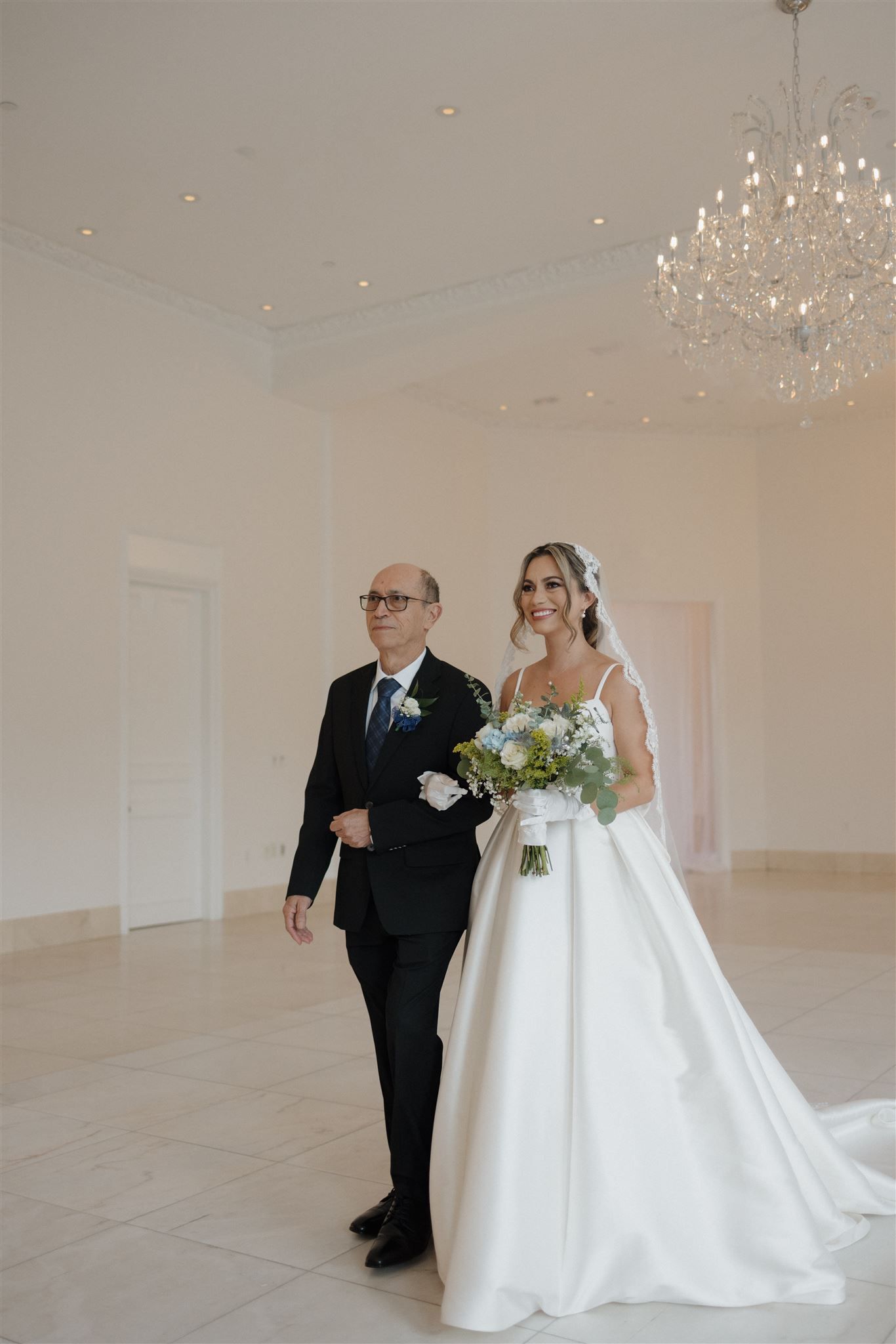 Two people stand together in an elegant white room with a crystal chandelier during a formal wedding ceremony.