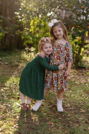 Two young children in dresses posing together outdoors during golden hour with autumn foliage in background.