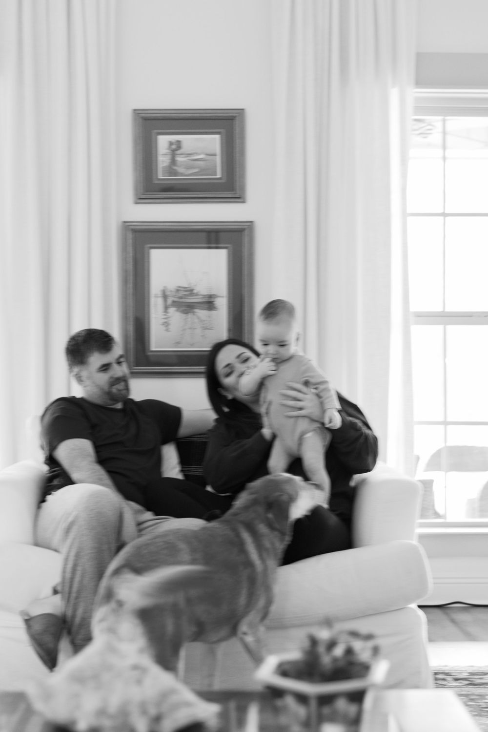 A black and white photo of family members relaxing on a white couch with two framed pictures hanging above.