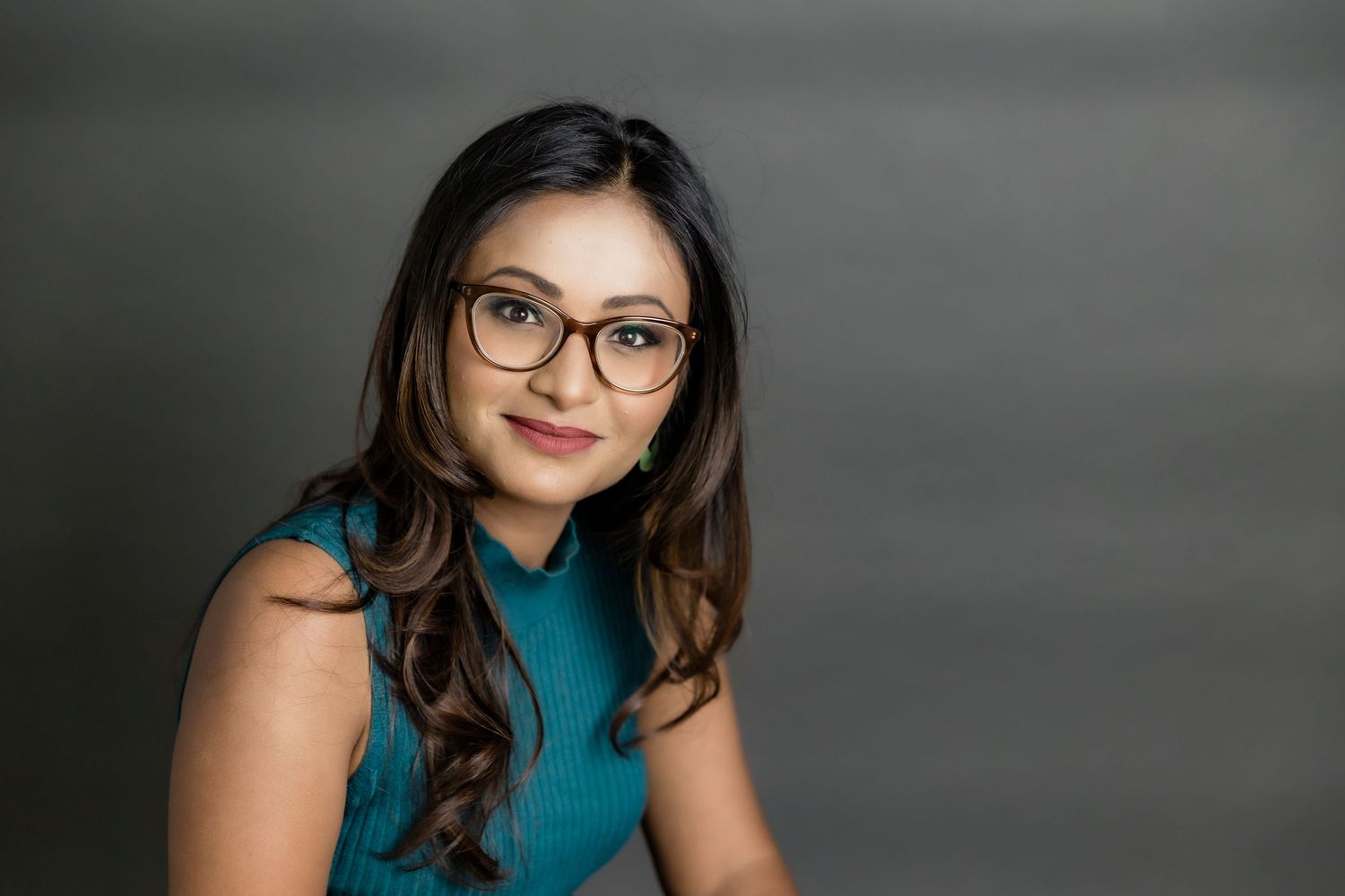 Professional portrait of a young and confident corporate consultant in a teal sleeveless top and glasses smiling against a gray background.