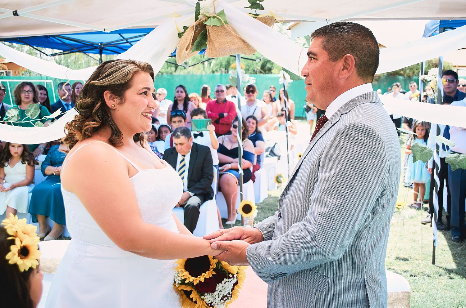 A couple exchanges vows during an outdoor summer wedding ceremony with sunflowers and white tent decor.