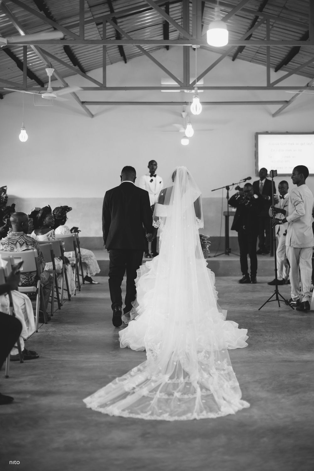 Black and white wedding ceremony photo showing bride's long train and veil in church setting.
