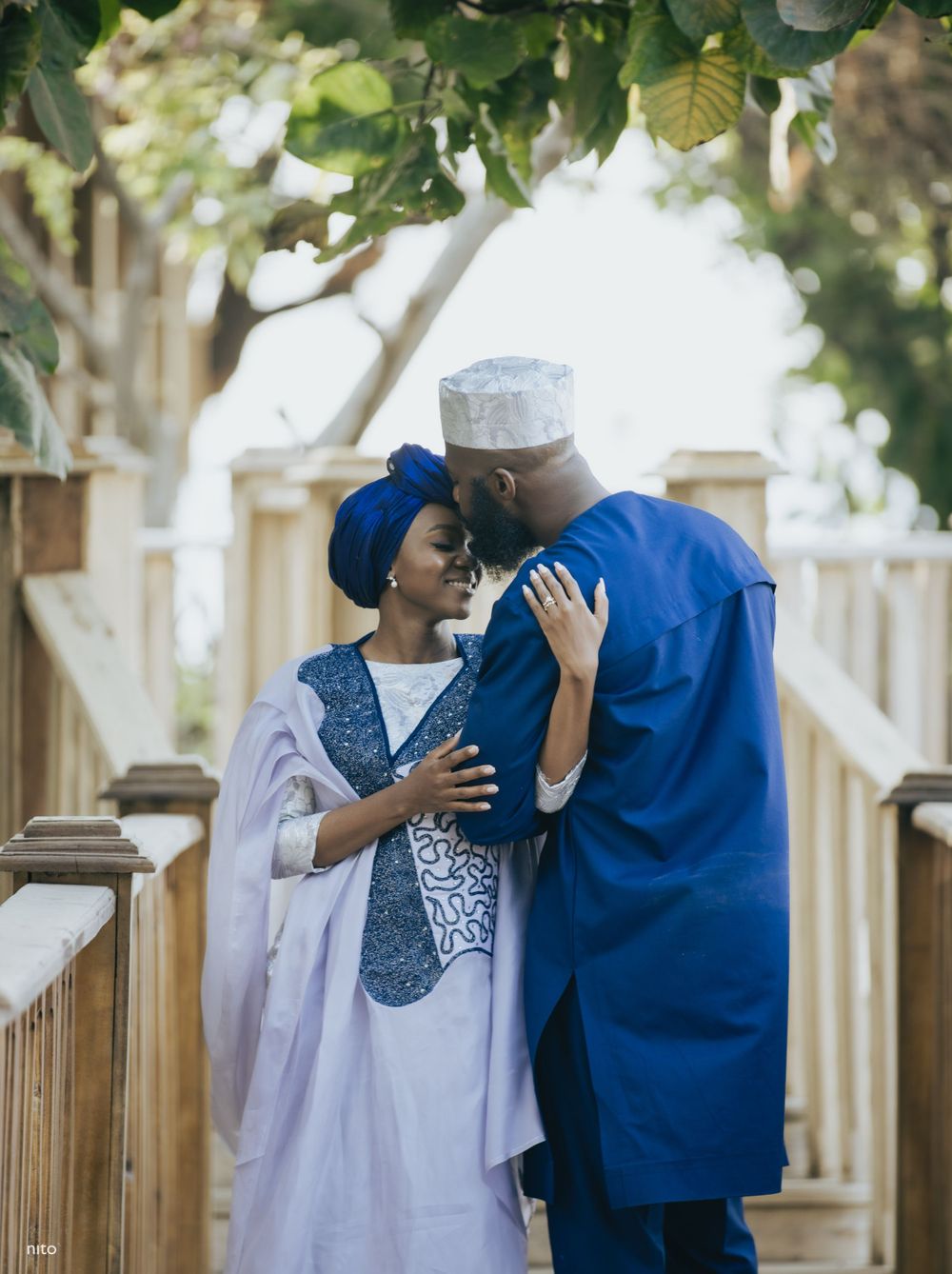 A couple shares an intimate moment on a white wooden bridge in traditional African dress.