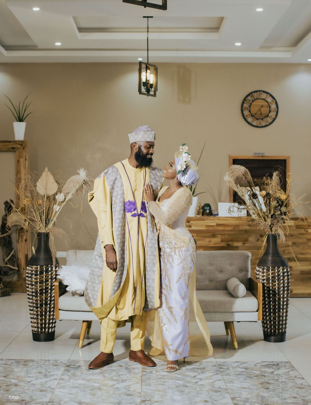 A couple in gold and white traditional attire poses in an elegantly decorated living room.