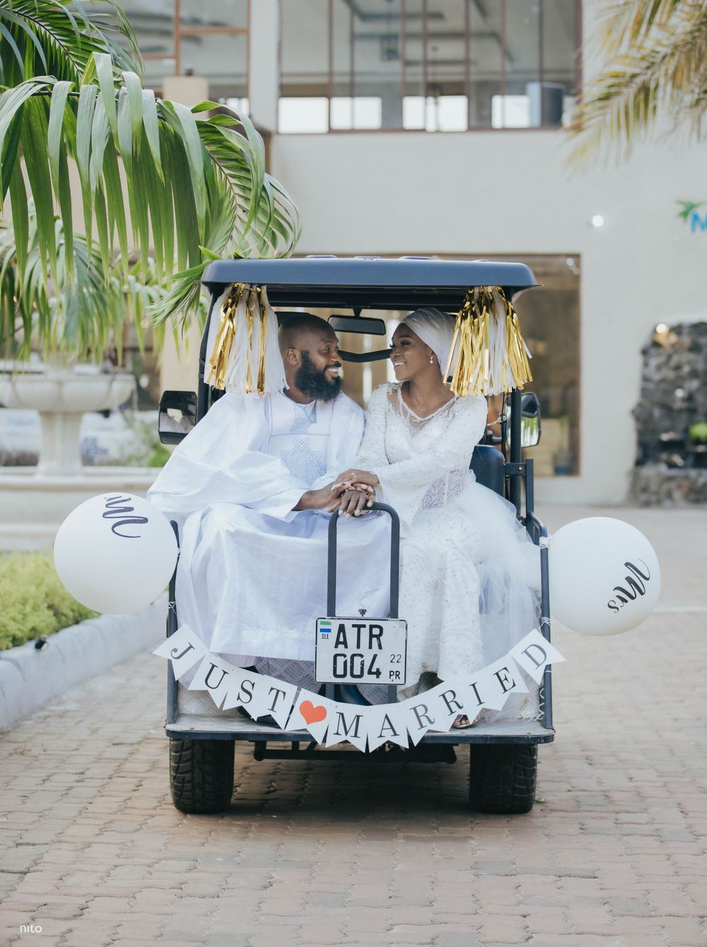 A newlywed couple sits in a decorated golf cart wearing matching white outfits.