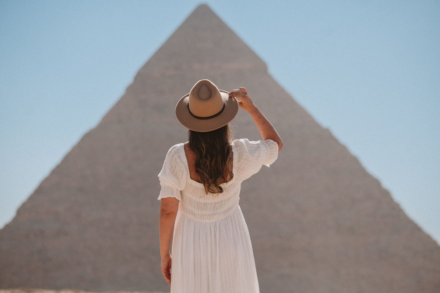 A tourist views the ancient pyramids of Giza in Egypt while wearing a flowing white dress and brown hat.