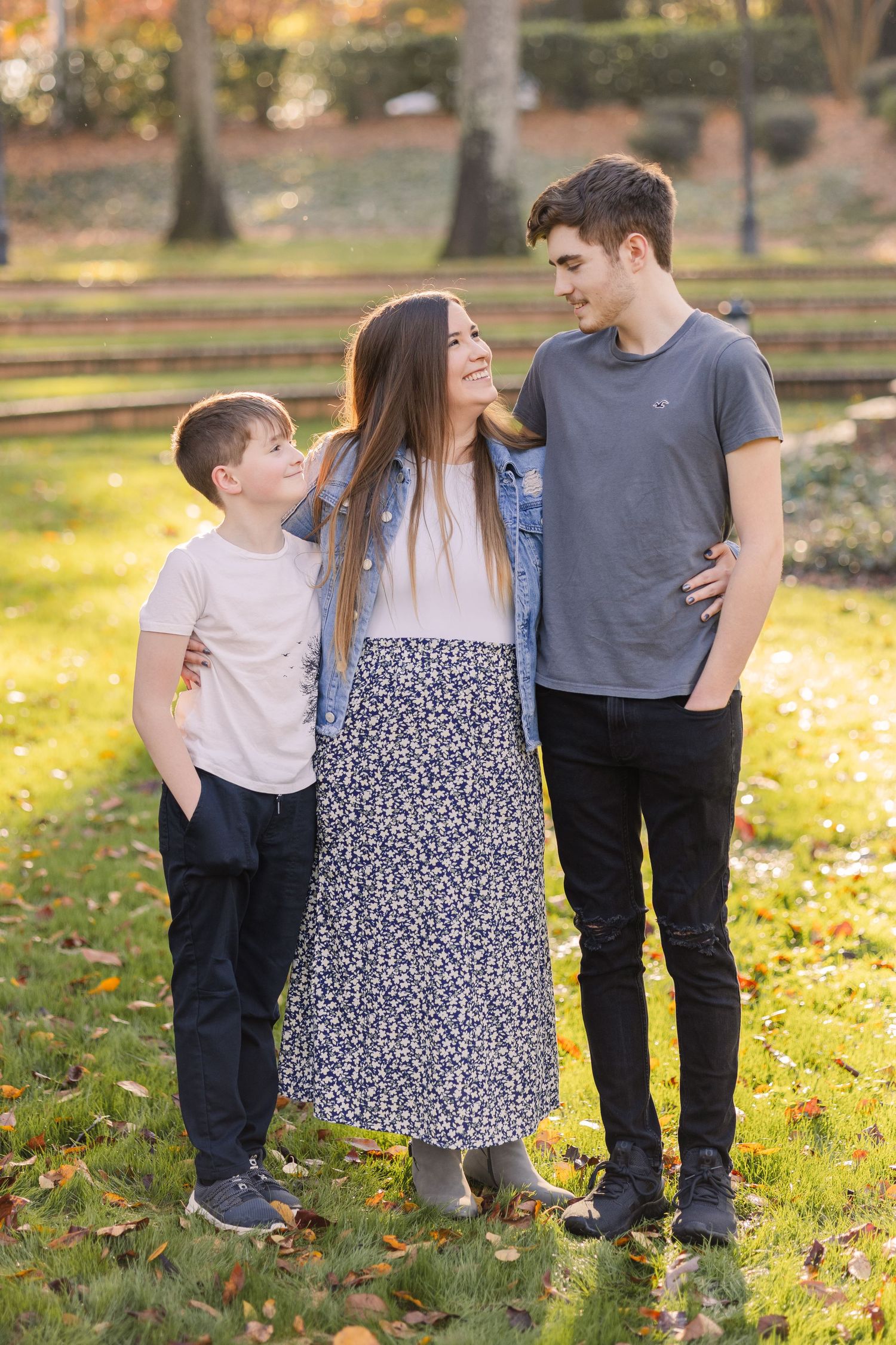Three people stand close together on fallen autumn leaves in a park setting during golden hour.