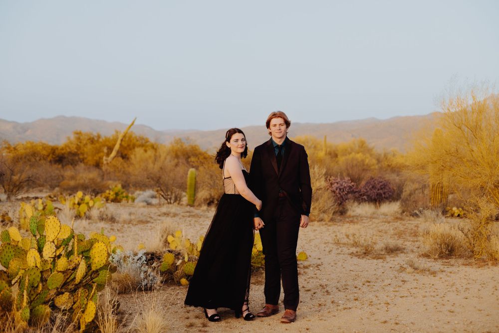 A couple poses together in dark formal wear against a desert landscape with cactus and mountains.
