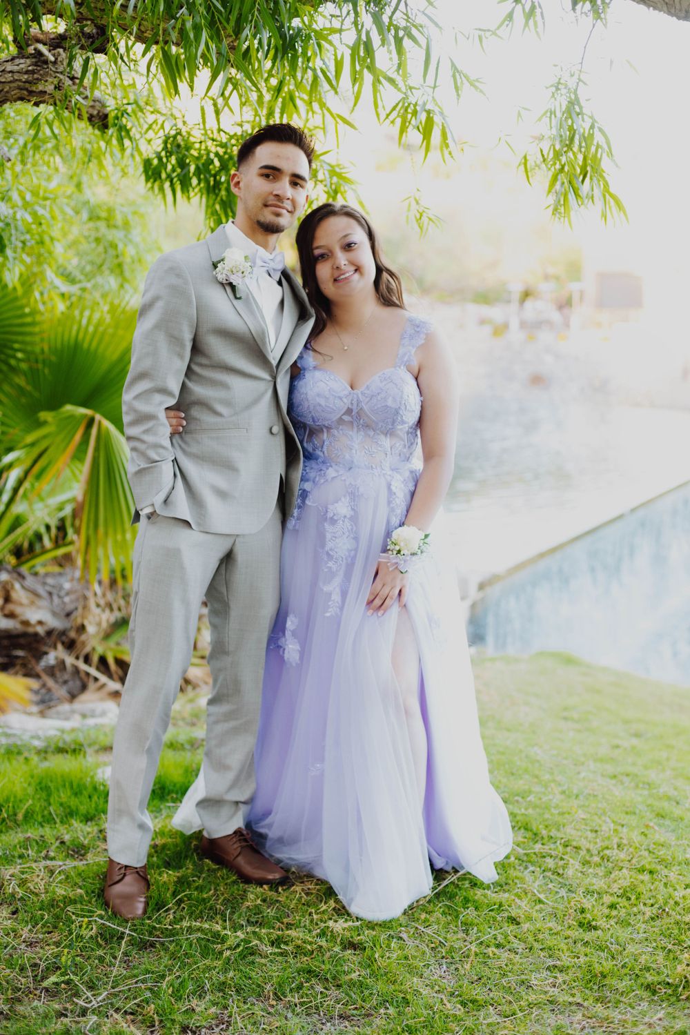 Couple in lavender dress and grey suit poses on grassy lawn with palm trees.