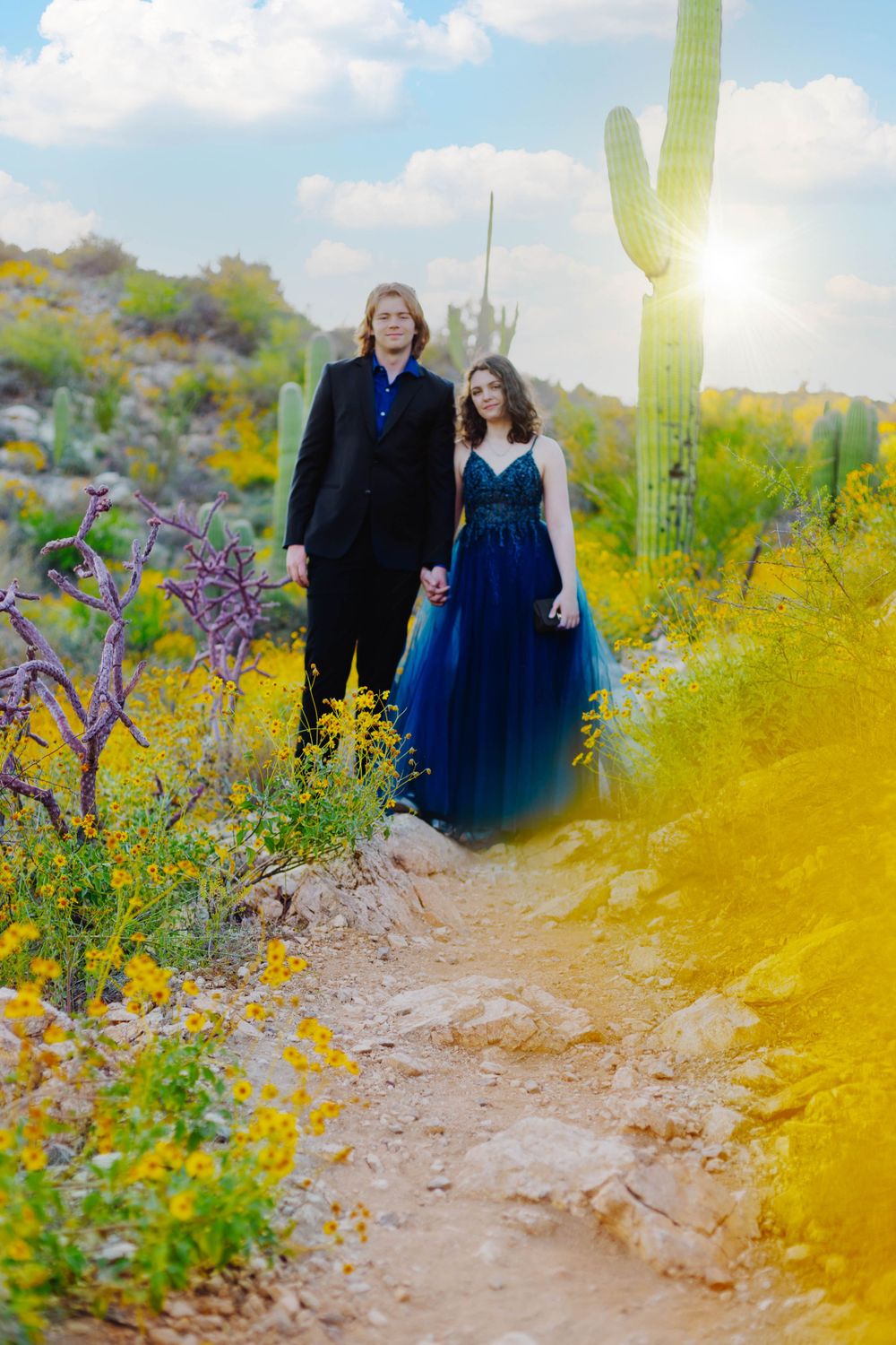 A couple in formal attire poses on a desert trail with yellow wildflowers and a tall saguaro cactus in the background.
