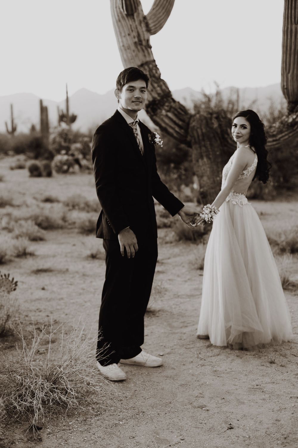 Black and white portrait of a couple in wedding attire posing beside a large saguaro cactus in the desert.