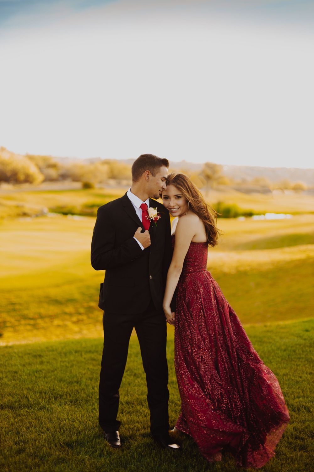 A couple in formal wear embraces on a grassy hill during golden hour sunset.