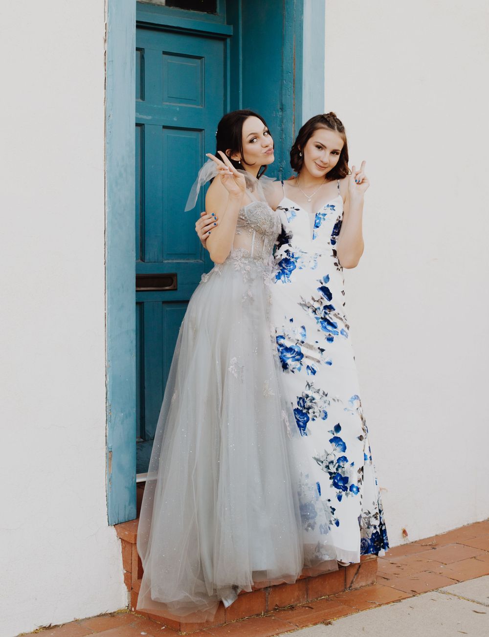 Two people wearing formal gowns pose in front of a turquoise door, one in grey tulle and one in blue floral.