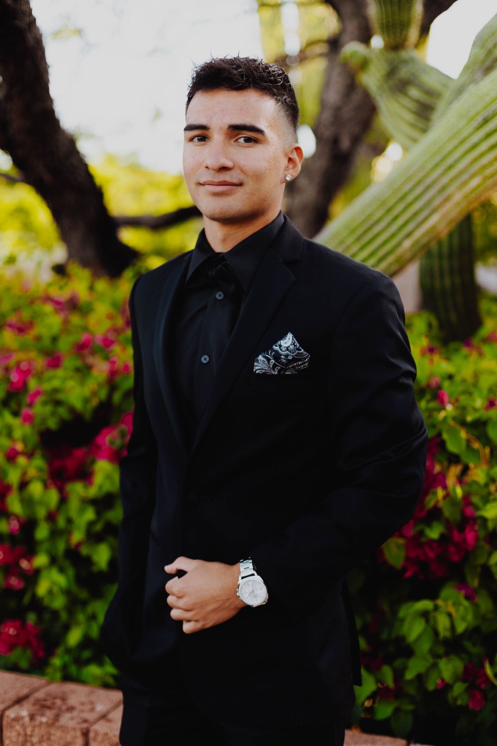 Man in black formal suit poses in garden setting with colorful flowers.