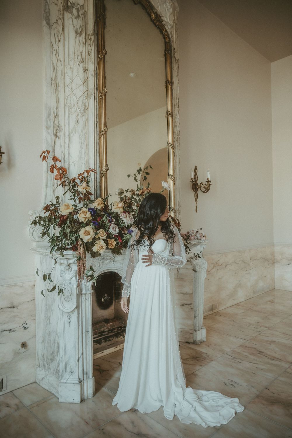 Elegant wedding dress silhouette against vintage fireplace with floral arrangement in ethereal light.
