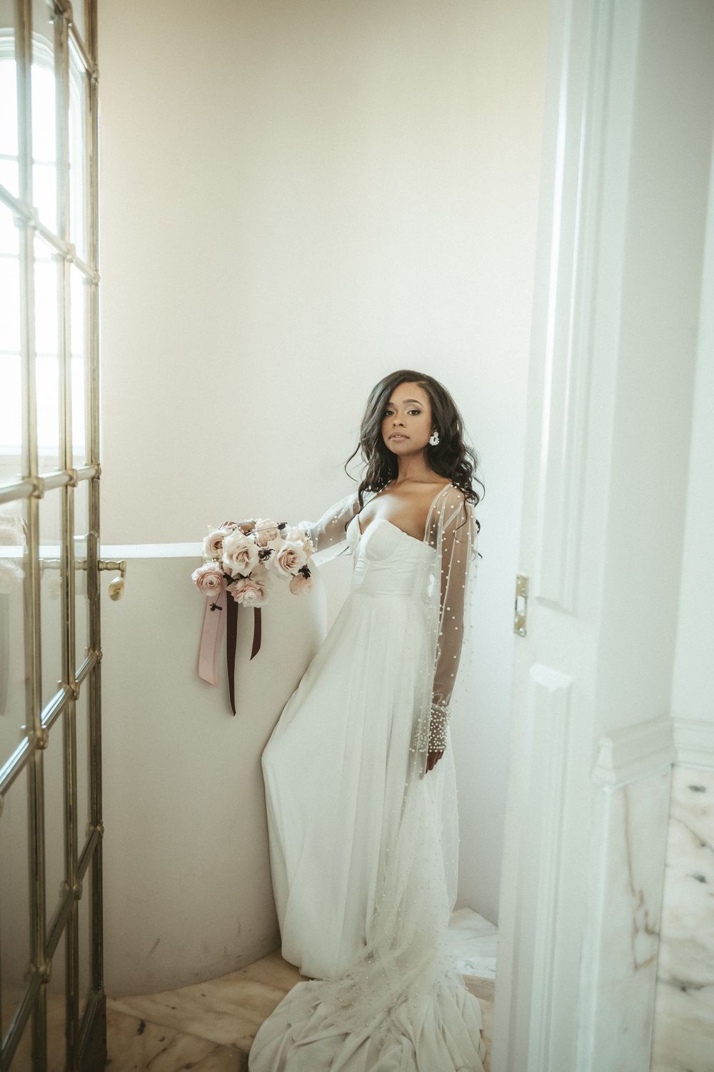 Elegant bridal portrait in flowing white gown with long sleeves against bright window.