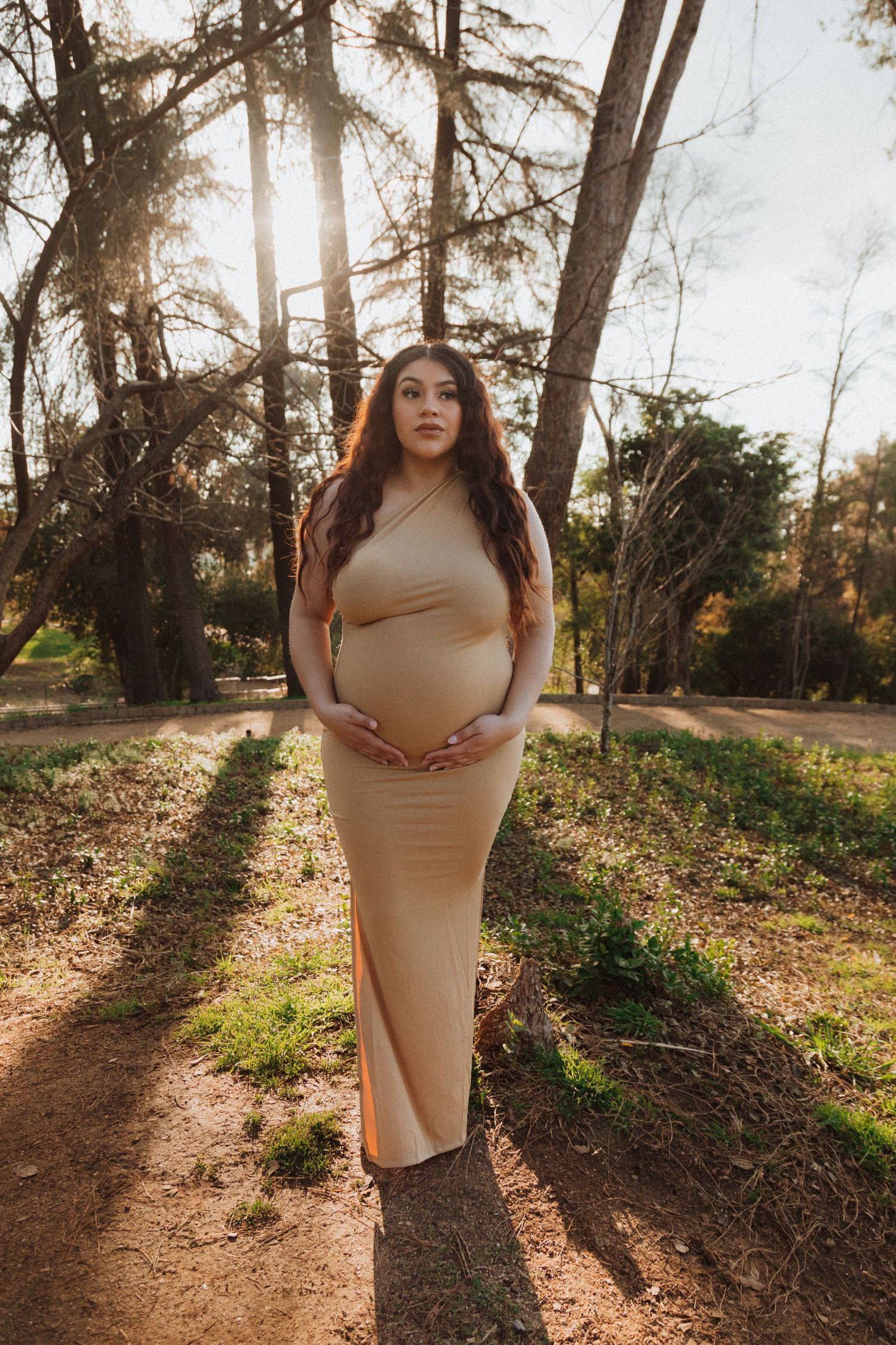 A maternity photo of a person in a tan dress standing in a sunlit wooded area.