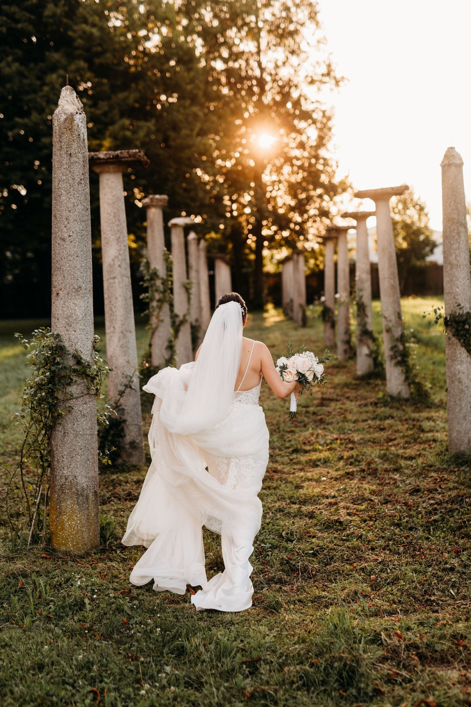 A bride in a white dress holding a bouquet poses among stone pillars at sunset.