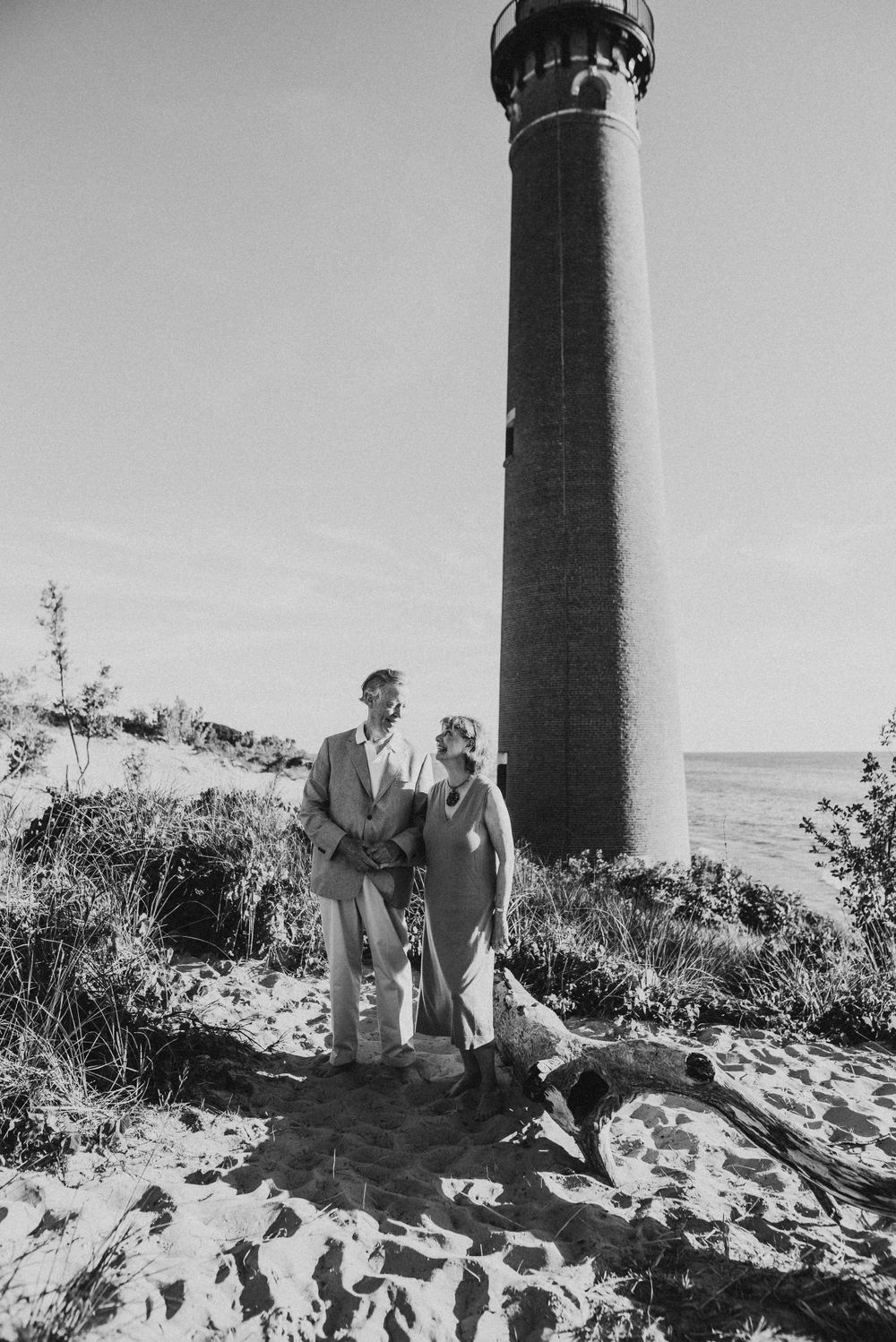 Black and white photo of a couple standing by a tall lighthouse on a beach.