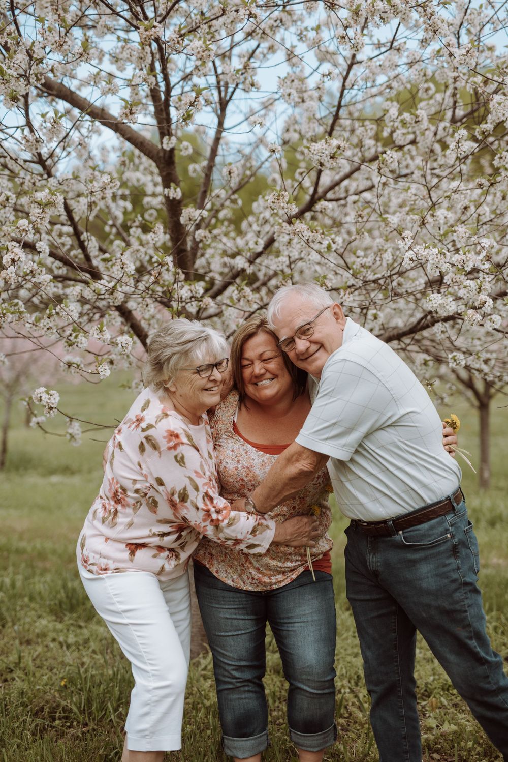 Three people sharing warm embrace outdoors under blooming cherry blossom trees.